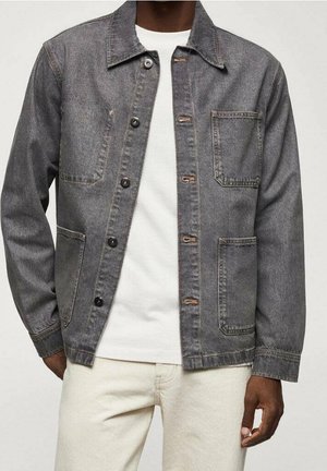 Homme portant une veste en denim grise avec des boutons noirs, une poche poitrine avant et deux poches carrées inférieures, sur une chemise blanche et un pantalon clair.