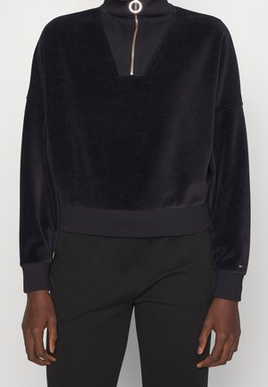 Persona che indossa un pullover nero in velluto a maniche lunghe con scollo a V a coste e dettaglio con cerniera dorata sopra pantaloni neri.