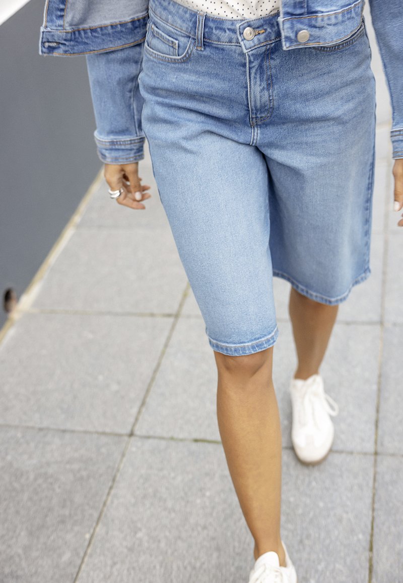 Personne marchant sur le trottoir portant un short en denim bleu clair, une veste en denim assortie, des baskets blanches et une chemise blanche à pois noirs.