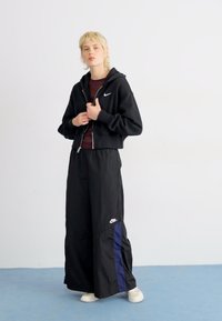 Hoodie noir cropped avec fermeture éclair, logo blanc, associé à un pantalon noir ample avec une bande latérale bleu marine et un logo blanc près de l'ourlet. Chaussures blanches.