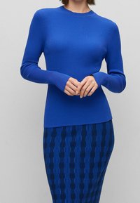 Pull bleu côtelé à manches longues avec une coupe ajustée et un col rond, associé à une jupe bleu foncé à motifs ornée de designs en forme de vagues texturées.