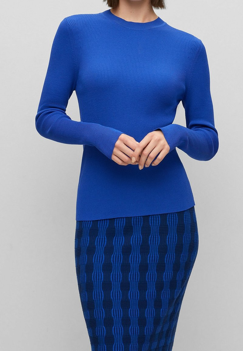 Pull bleu côtelé à manches longues avec une coupe ajustée et un col rond, associé à une jupe bleu foncé à motifs ornée de designs en forme de vagues texturées.