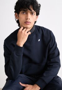 Felpa girocollo blu navy con tessuto testurizzato, polsini a coste e un piccolo logo bianco sul petto. Il modello posa con una mano sul mento.