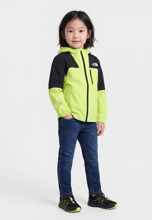 Dieťa oblečené v neonovo žltej a čiernej bunde, modrých džínsoch a čiernych teniskách s žltými akcentmi, stojace s jednou rukou v vrecku.