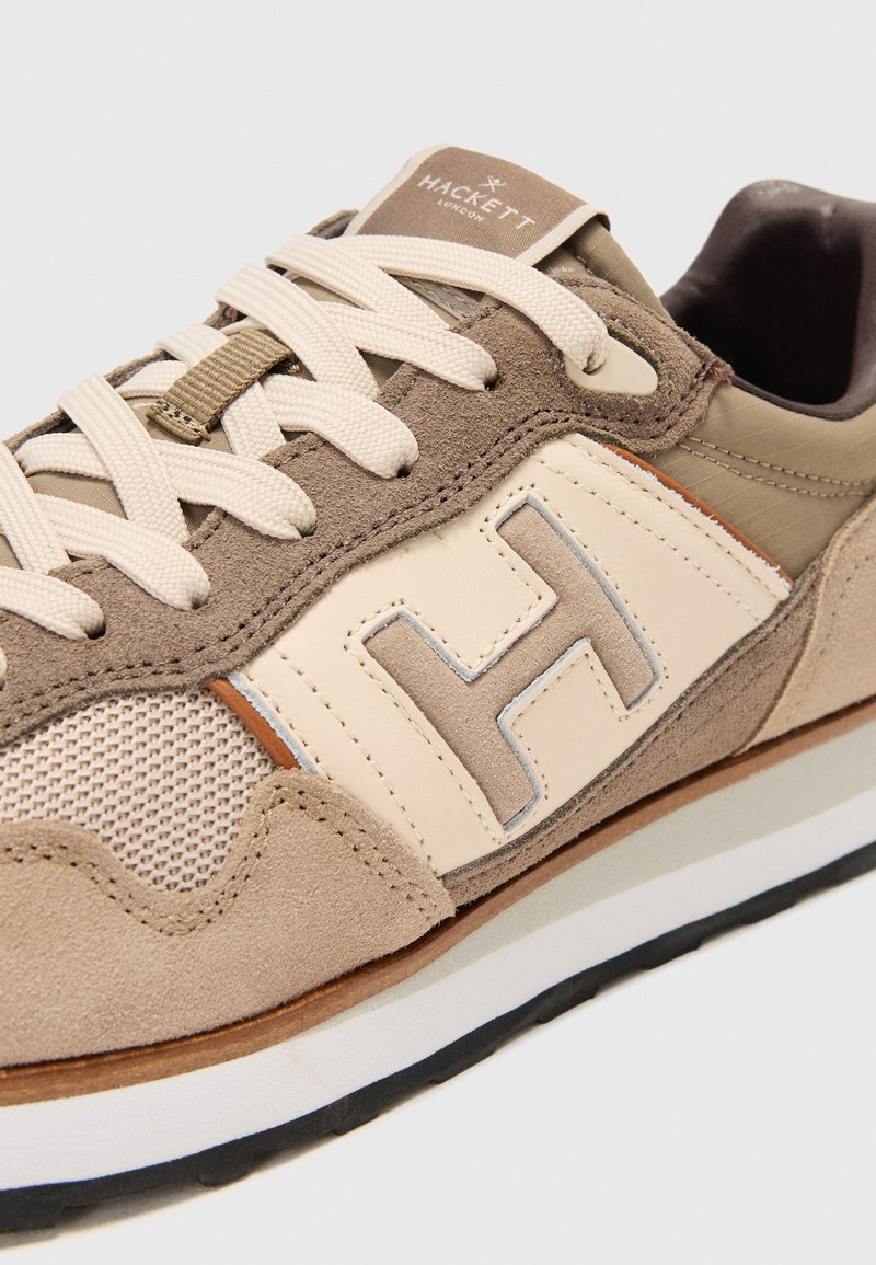 Gros plan sur une sneaker Hackett London beige et marron avec des détails en daim, des panneaux en maille, des lacets crème et un grand "H" brodé sur le côté.