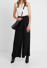 Biała bluzka z dekoltem w kształcie kokardy, czarne szerokie spodnie oraz czarne botki na obcasie z ostrym noskiem. Czerwona torebka na ramię dopełnia stylizację.