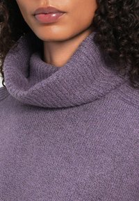 Nahaufnahme einer Person, die einen strukturierten, lavendelfarbenen Rollkragenpullover trägt. Zu sehen sind Lippen, Kinn und lockiges, dunkles Haar.