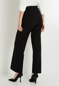 Mujer con pantalones negros de tiro alto y ajustados, y zapatos de tacón negro, vista desde la espalda contra un fondo liso.