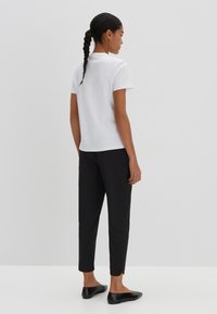 Weißes kurzärmliges Oberteil kombiniert mit schwarzen tapered Hosen. Schwarze flache Schuhe haben einen kleinen Akzent. Der Hintergrund ist einfarbig und hell.