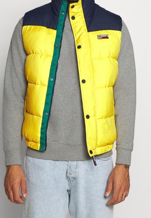 Mężczyzna ubrany w żółto-granatową pikowaną kamizelkę na szarą bluzę i jasnoniebieskie jeansy, stojący na neutralnym tle.