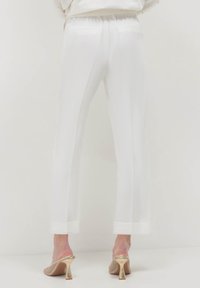 Persona vestita con pantaloni bianchi su misura con orli risvoltati e mule con tacco alto in rete dorata, in piedi contro uno sfondo bianco uniforme.