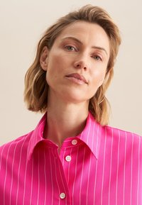 Fuchsia overhemd met knopen en witte verticale strepen, met een puntige kraag en een gladde textuur, gepresenteerd tegen een neutrale achtergrond.