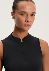 Fekete ujjatlan felső magas gallérral és elülső cipzárral, sima anyagból készült. Fitt szabású és minimális részletezéssel rendelkezik.