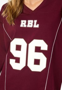 Bordo pulover sa V-izrezom, bijelim detaljima, tekstom "RBL" i velikim brojem "96" u bijeloj boji s bež obrubom. Mekana tkanina.
