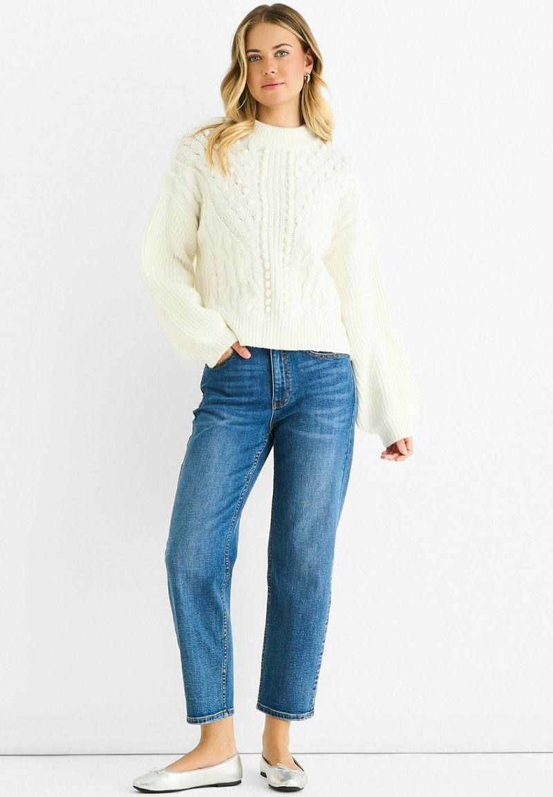 Cream cable-knit sweater with ribbed cuffs, paired with straight-leg blue jeans and silver ballet flats.