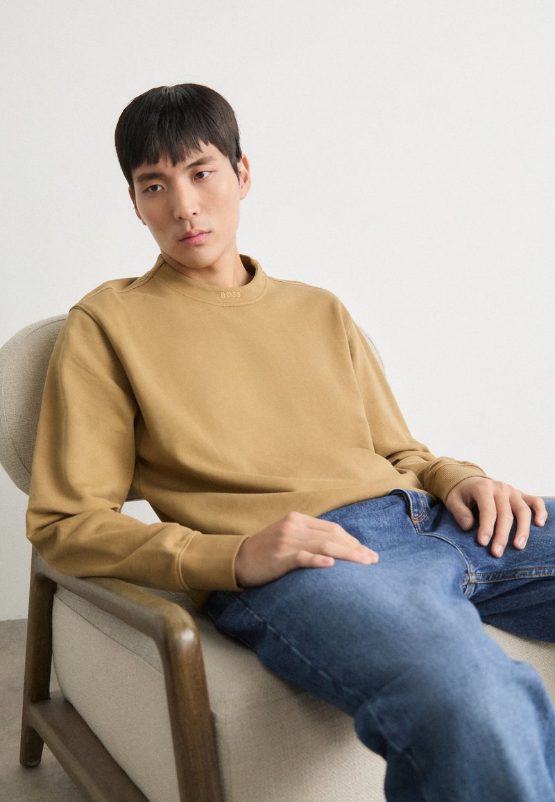 Sudadera marrón con cuello redondo y detalles discretos del logo, combinada con jeans azules. Modelo sentado en una silla de color claro. Fondo minimalista.