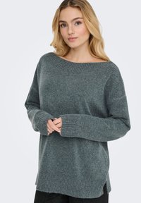 Siv pulover iz pletenine z širokim izrezom, dolgimi rokavi, rebrastimi zapestji, stranskimi režami in mehko teksturo. Prijeten in sproščen kroj, sega pod boke.