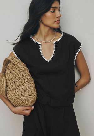 Žena s dlhými čiernymi vlasmi, ktorá nosí čierne tielko s bielym lemom, drží pletenú slamennú tašku a nosí perlový náhrdelník a zlaté kruhové náušnice.