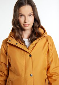 Gele outdoorjas met capuchon, voorzien van drukknoopsluiting, trekkoordaccenten en een gladde textuur. De stof lijkt duurzaam en weerbestendig.