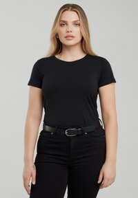 Jonge vrouw met lang blond haar, gekleed in een zwart shirt met korte mouwen, zwarte spijkerbroek en een zwarte riem, staand tegen een grijze achtergrond.