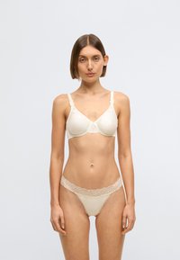 Sutiã bege e tanga combinando com detalhes em renda. O sutiã tem alças ajustáveis e um design liso e ajustado. Detalhe em renda texturada ao redor da cintura.