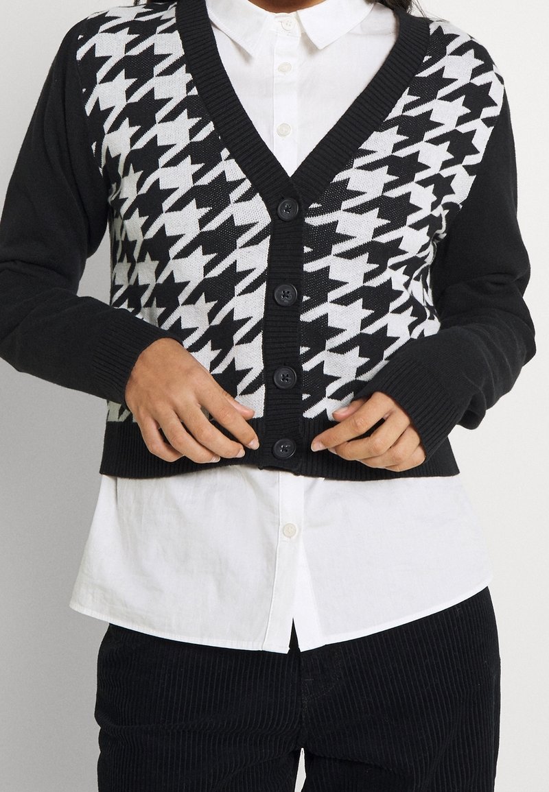 Personne boutonnant un cardigan pied-de-poule noir et blanc par-dessus une chemise blanche avec un pantalon noir, la taille visible sur un fond uni.