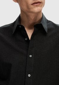 Camicia nera con colletto classico, caratterizzata da una texture morbida e una leggera lucentezza. Tre bottoni visibili sul collo.