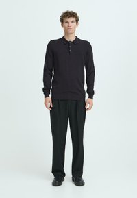 Mørk strikket polo trøje med en tre-knap lukning, båret med sorte brede bukser og sorte sko, mod en lys baggrund.