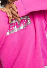 Sudadera de un brillante color rosa con un logo rectangular plateado metálico que lleva la palabra "REPLAY" en letras negritas en el pecho.