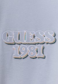 Tela azul claro con el logotipo "GUESS" y "1981" en bordado 3D, con múltiples acentos de color, incluyendo contornos en blanco, naranja y verde azulado.