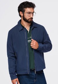 Homme aux cheveux bouclés et à la barbe portant des lunettes, une veste zippée bleue, un polo vert et un jean en denim sur un fond uni.