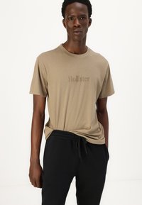 Taupe kokvilnas T-krekls ar īsām piedurknēm, apaļu apkakli un izspiedu "Hollister" logo uz priekšpuses. Kombinēts ar melniem elastīgiem sporta biksēm.
