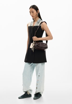 Vrouw draagt een mouwloos zwart blazer met denim kraag, lichte jeans met wijde pijpen en zijknopen, zwarte sneakers, en houdt een donkerbruine schoudertas vast.