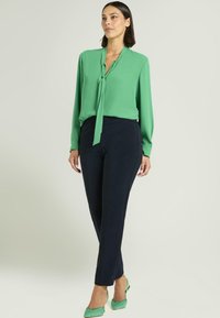 Groene blouse met lange mouwen en strikdetail, gecombineerd met donkerblauwe nauwsluitende broek. Model draagt groene puntige platte schoenen. Gladde stof, getailleerde pasvorm.