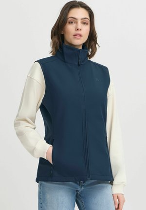 Gilet sans manches bleu marine avec col montant, fermeture éclair et poches latérales ; manches longues blanches visibles en dessous ; tissu lisse et léger.