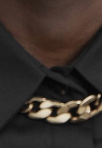 Schwarzes Hemd mit Kragen und einer goldenen Kettenhalskette. Die Kette hat große, miteinander verbundene Glieder mit einem glänzenden Finish.