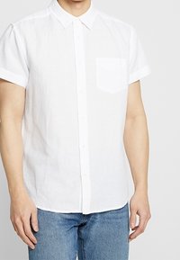 Camicia bianca a maniche corte con bottoni, in lino, con taschino a petto e colletto classico. Indossata con jeans denim blu.