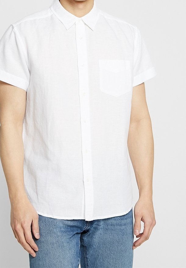 Camicia bianca a maniche corte con bottoni, in lino, con taschino a petto e colletto classico. Indossata con jeans denim blu.