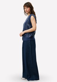 Haut sans manches bleu tricoté avec un motif texturé, associé à un pantalon large en velours côtelé bleu marine. La tenue présente une silhouette décontractée.