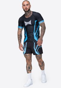 Melnā sporta krekls ar zilajiem akcentiem un izsmidzinājuma rakstu; apvienots ar melniem šortiem, kuru dizainā ir zilas ģeometriskas formas. Abas drēbes ir ar zīmolu.