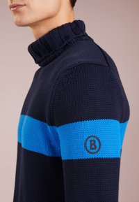 Maglione a maglia blu navy con colletto alto, caratterizzato da una striscia orizzontale blu e un logo sulla manica. Tessuto testurizzato con dettagli a coste.