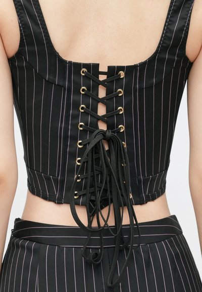 Top corto negro con rayas verticales, que presenta un diseño de espalda con cordones, con ojales dorados y un cordón negro. Textura suave.