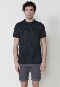 Crna polo majica kratkih rukava s plavim geometrijskim uzorkom. Ima ovratnik s dugmadi i glatku tkaninu. Uparena je s sivo-rolanim kratkim hlačama.