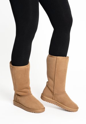 Beine mit schwarzen Leggings und mittellangen tanfarbenen Wildleder-Stiefeln mit flachen, gemusterten Sohlen auf weißem Hintergrund.