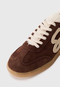 Brown suede sneaker with cream laces, shearling lining, and a light brown rubber sole. Features an embossed logo on the side.