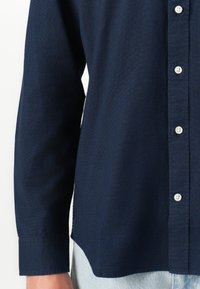 Camisa de botones de manga larga azul marino usada sobre unos vaqueros azul claro, mostrando detalles de los puños, botones y la costura lateral.