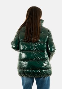 Chaqueta acolchada verde brillante con un cuello alto, acolchado horizontal y un corte holgado. Combinada con jeans de mezclilla azules.