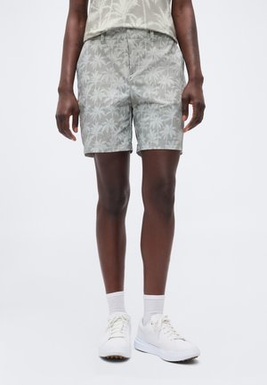 Person trägt graue Shorts und ein passendes Hemd mit Palmenmuster, weiße Socken und weiße Turnschuhe, steht vor einem einfarbigen Hintergrund.