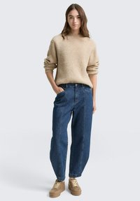 Beiger gerippter Pullover mit weiten Ärmeln, kombiniert mit hochgeschnittenen dunkelblauen Jeans und braunen Plateau-Sneakern. Einfacher, lässiger Schnitt.