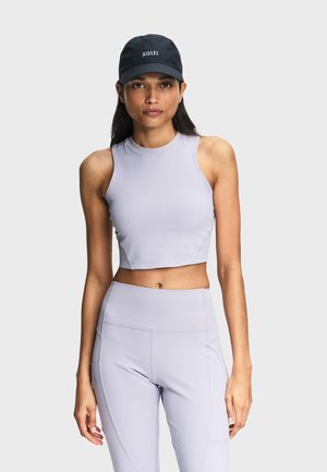 Femme portant une casquette noire et un haut court sans manches gris clair, avec un legging taille haute assorti, sur un fond uni.